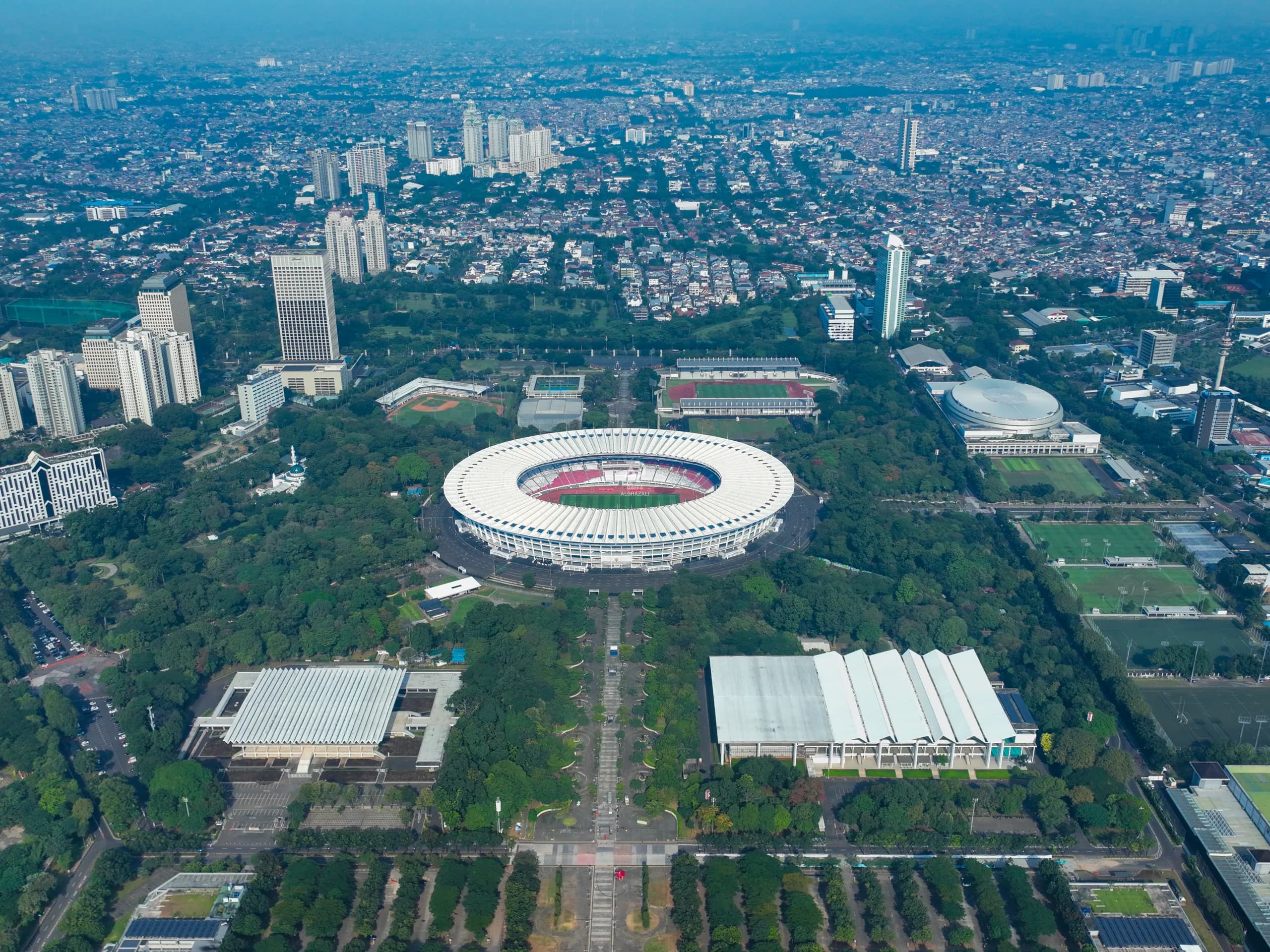 An aerial perspective of Gelora Bung Karno (GBK), showcasing its iconic stadium, surrounding sports facilities, and expansive green spaces in the heart of Jakarta. This wide-angle composition captures the scale of the sports complex and its seamless integration with the city’s urban landscape, highlighting the architectural beauty and vibrant character of Indonesia’s capital.